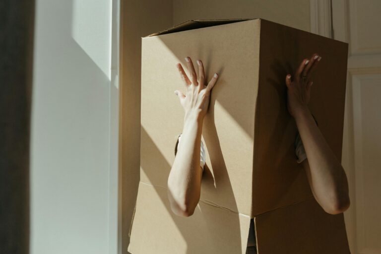 A person hides inside a large cardboard box, playfully showing their hands.